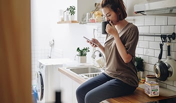 Woman sitting on kitchen counter looking at phone drinking coffee