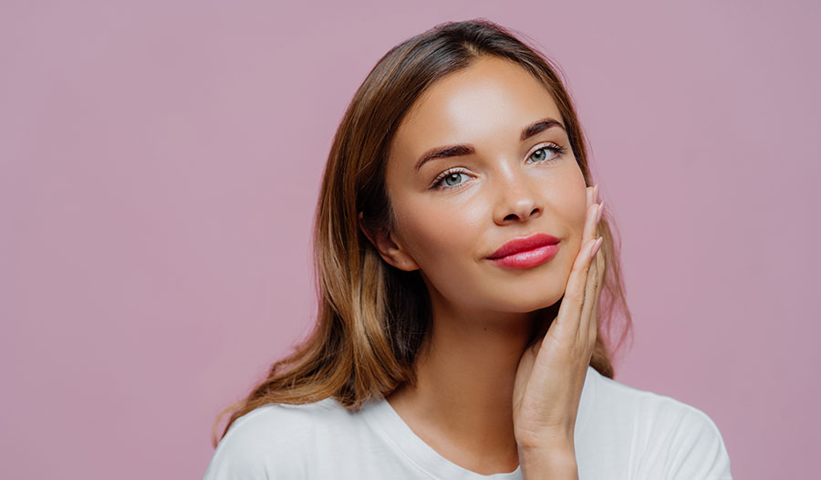 woman with glowing skin touching her face with one hand against a mauve background