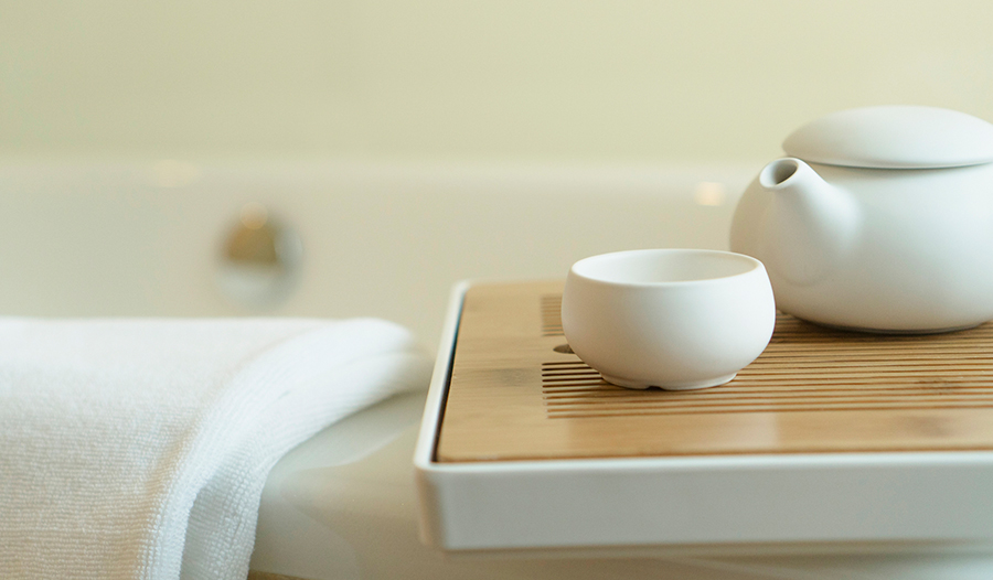 white teapot and teacup on a wooden tray next to a towel resting on the edge of a bathtub