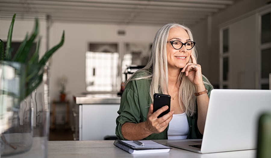 Happy senior woman with smartphone and laptop working from home 