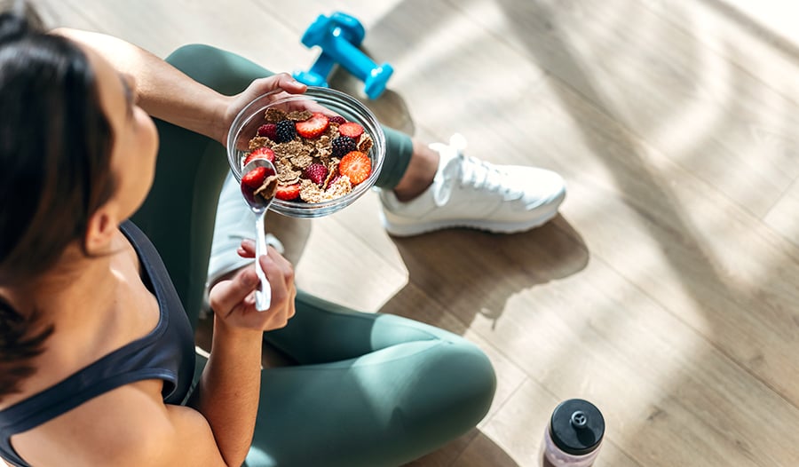 Healthy woman eating bowl of cereal and fruit with weights and water bottle