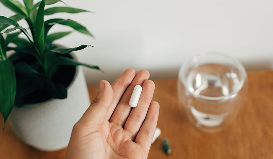 Hand holding supplement in hand above table with glass of water and plant