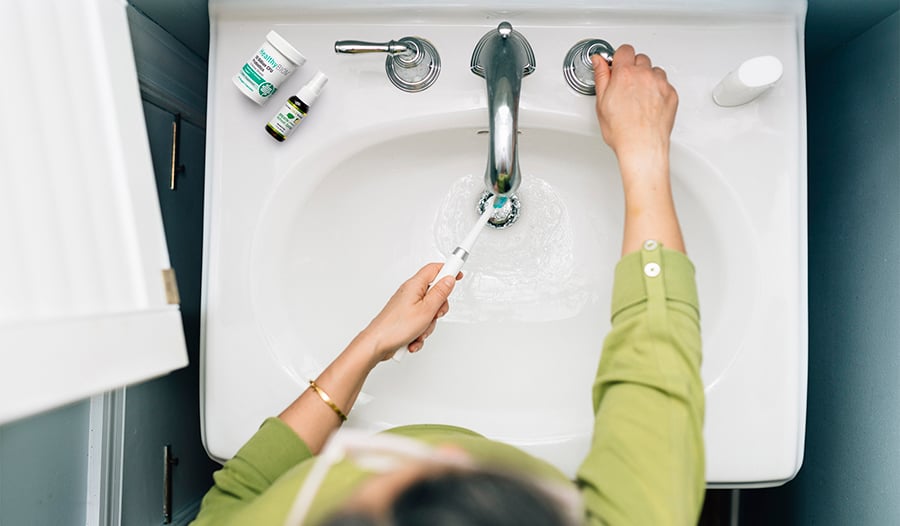 Person brushing teeth at bathroom sink with probiotics and breath spray on counter