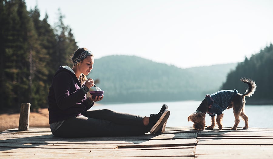 Woman eating healthy food on wooden pier around mountain lake with her dog.