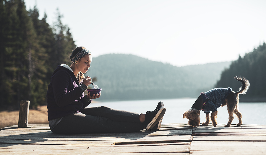 Woman eating healthy food on wooden pier around mountain lake with her dog.