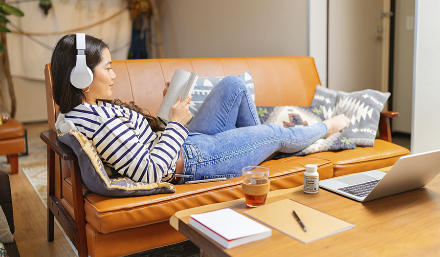 Woman with headphones on laying on couch reading with tea and digestive enzymes on table