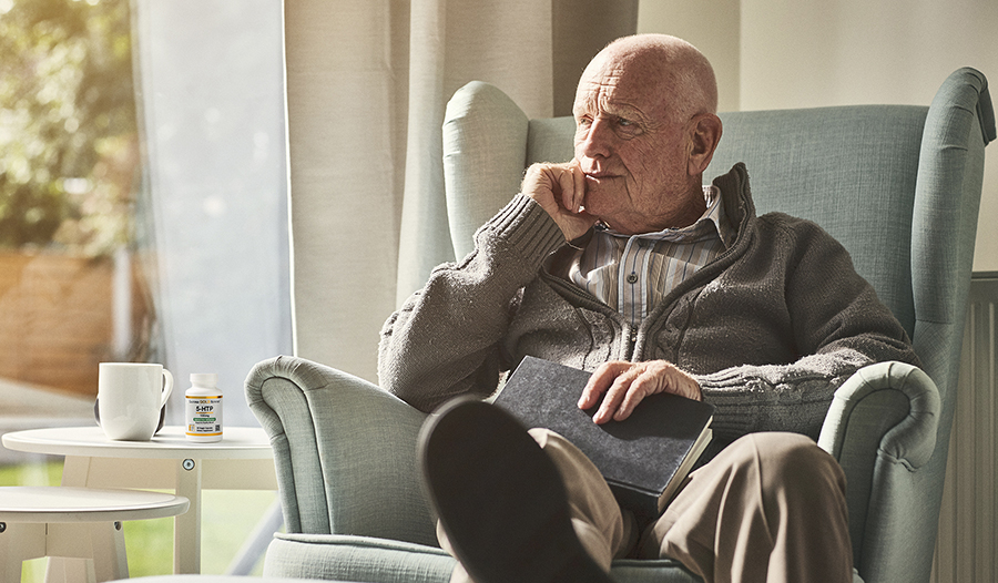 Mature male sitting in chair by window with tea and 5-HTP supplement on table