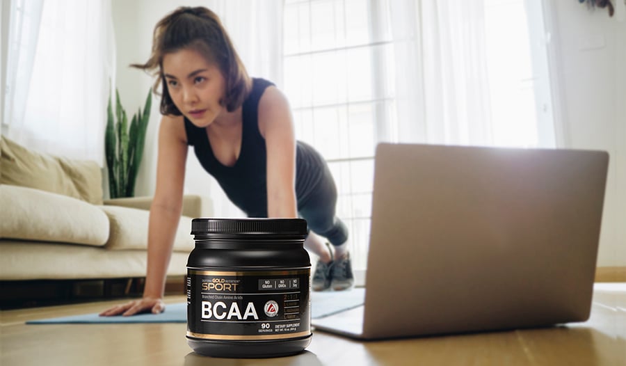 Woman working out at home with laptop in living room; BCAAs supplement in foreground