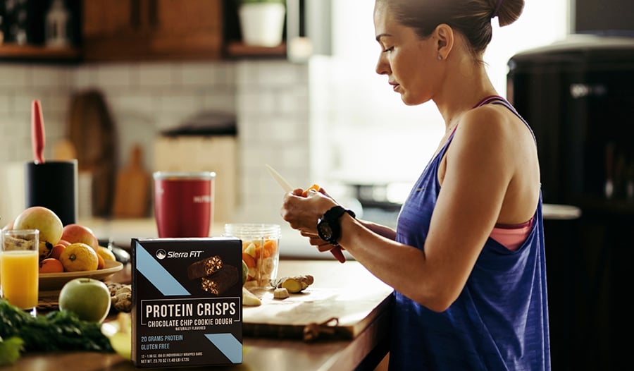 Une femme préparant un smoothie dans sa cuisine avec des fruits frais et des snacks protéinés.